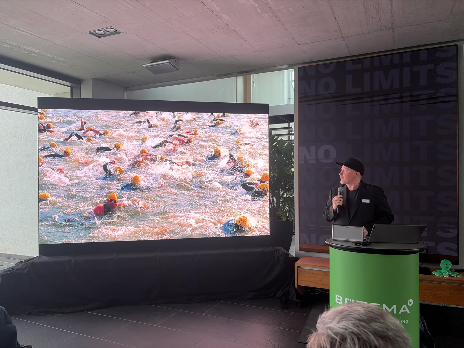Joey Kelly auf der Bühne, auf der Leinwand ein Feld von Schwimmern beim Start eines Wettkampfs